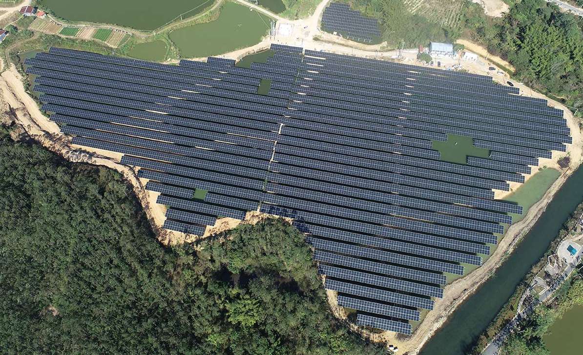 Aerial view of a large-scale ground-mounted solar PV plant surrounded by forest and ponds, showcasing utility-scale solar deployment in a rural area